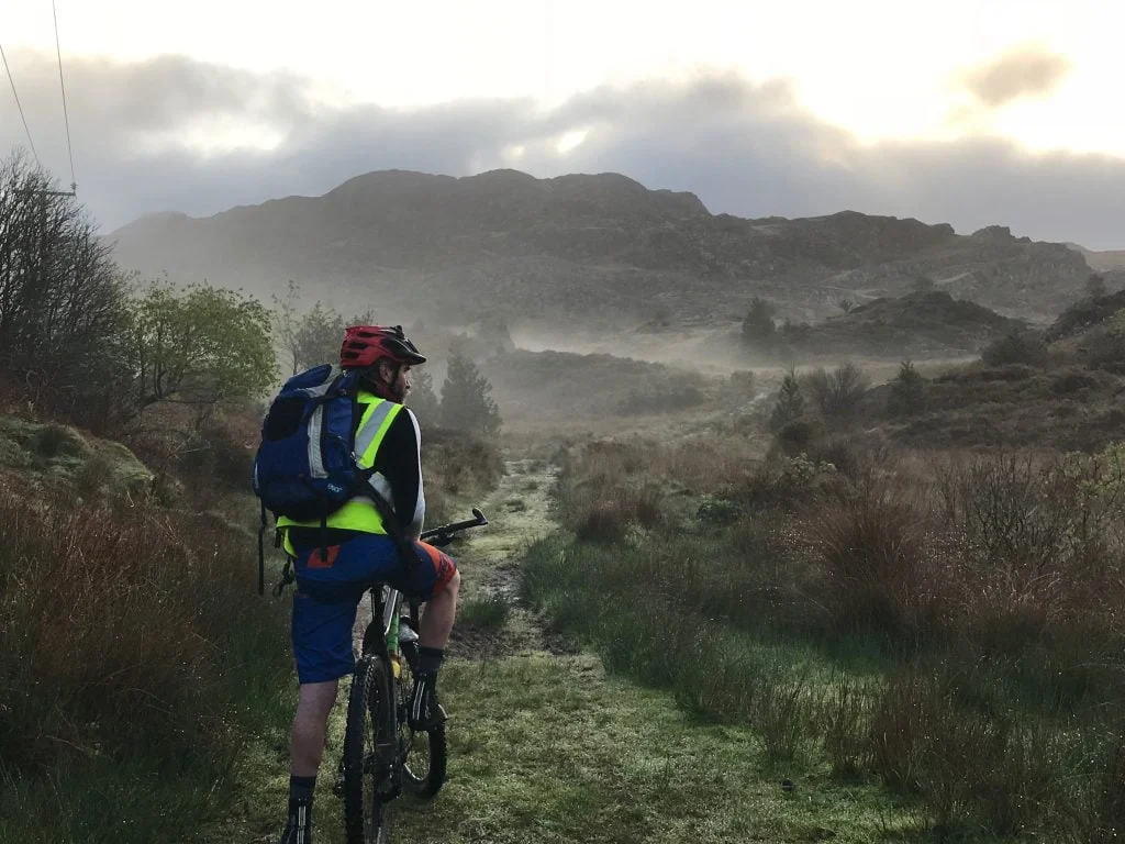 Scot Tares of Skinny Tyres stops to admire the beautiful scenery as he rides a mountain bike across a grassy, shrubby area towards a rocky hill. The grass is clearly wet and the early morning mist can be seen in the background.