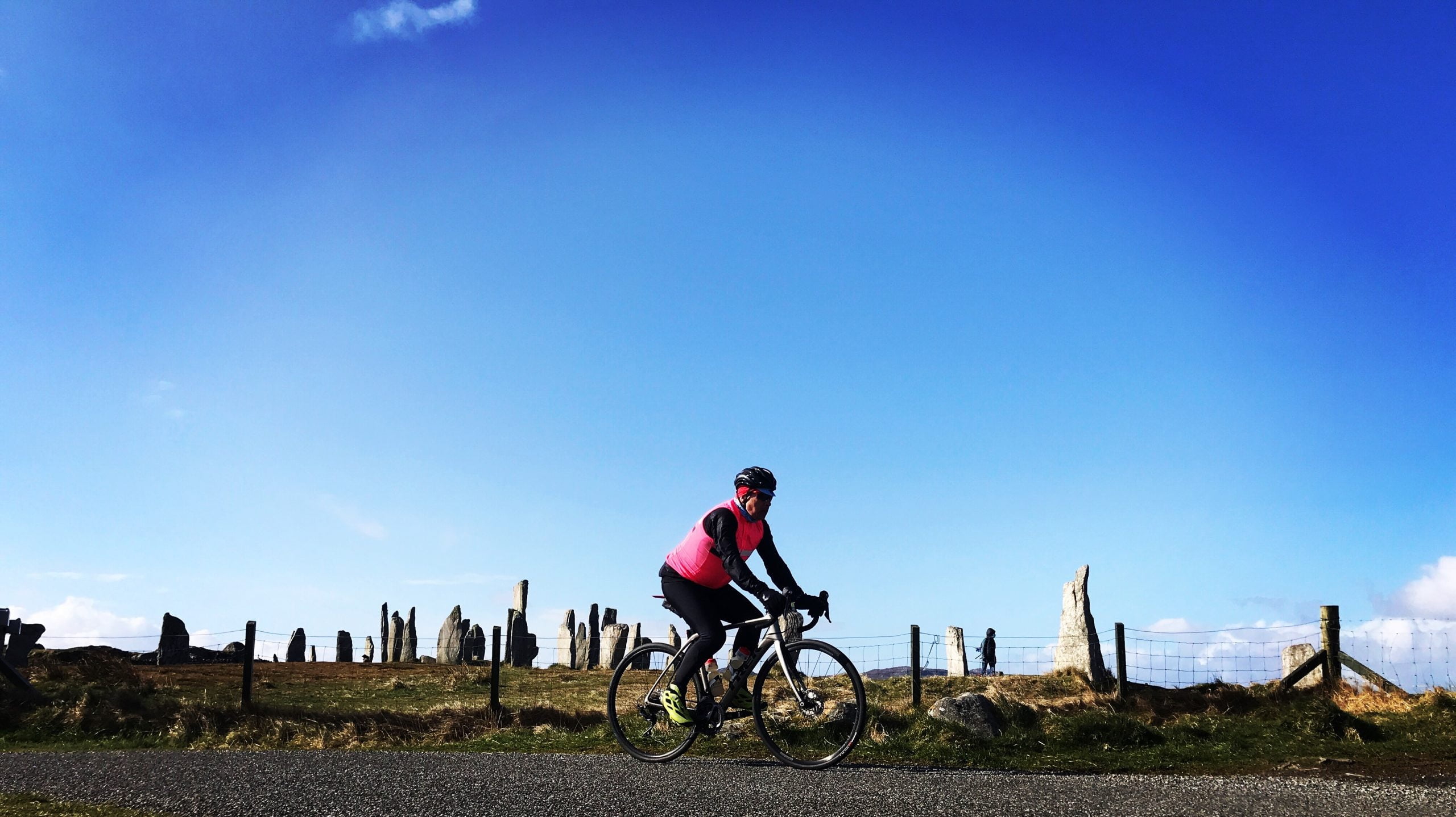 A cyclist in cold weather gear and a red gilet cycles past the Callanish Standing Stones on the Isle of Lewis on a guided road cycling holiday with Skinny Tyres Ltd.