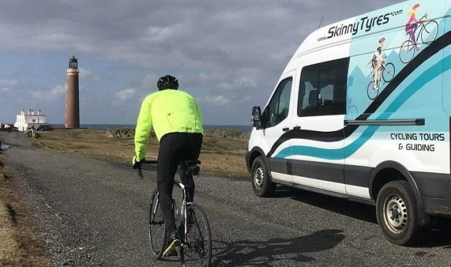 A solo cyclist in bright winter gear on a Skinny Tyres guided road cycling holiday in Scotland passes the fully customised support van just before the Stevenson Lighthouse at the Butt of Lewis on the Hebridean Way.