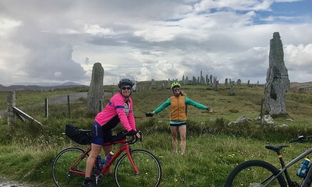 Two female cyclists wearing colourful jackets on a Skinny Tyres Cycling Tour pose with their bikes at the Callanish Standing Stones on Lewis.
