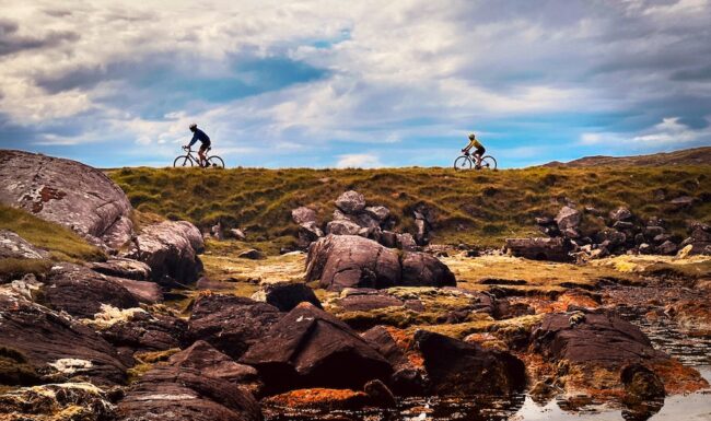 Two cyclists on the horizon riding on the Golden Road, Harris, Outer Hebrides as part of the Hebridean Way cycle tour Cycling in Scotland.