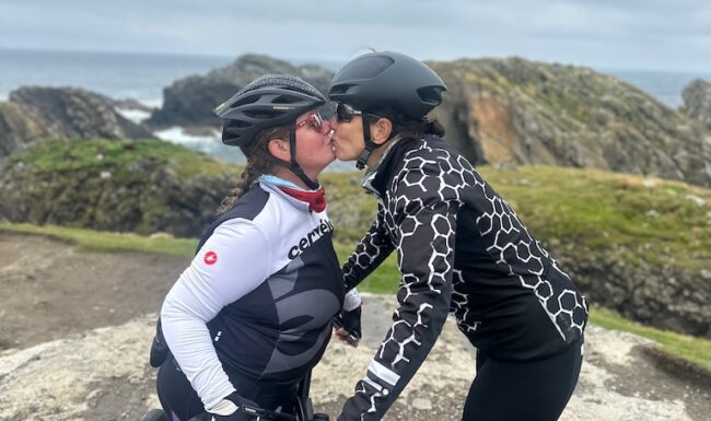 An LGBTQ female couple on a Skinny Tyres cycling holiday in Scotland kiss at the Butt of Lewis, congratulating each other for reaching the end of their Hebridean Way trip. The Atlantic Ocean can be seen beyond the rocks in the background.