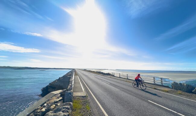 A cyclist rides on the Hebridean Way. She is cycling across a causeway that links Benbecual to North Uist. Behind her the sky is blue and the sun is blazing in the sky The image was taken on a Skinny Tyres Tour Cycling Holiday The Hebridean Way.