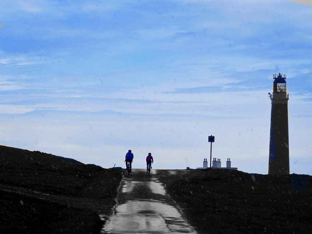 A man and a woman cycle towards the end of the hebridean Way. They are silhoutted against a blue sky and a lighthouse is on the horizon to their right.