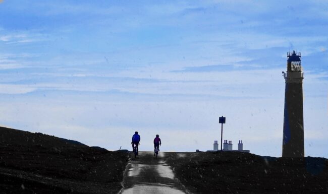 A man and a woman cycle towards the end of the hebridean Way. They are silhoutted against a blue sky and a lighthouse is on the horizon to their right.