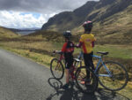 ​A young boy and girl stand astride their bikes looking south towards Lochan na Lairige, just below the summit of Ben Lawers. The girl is wearing a Soreen Malt Loaf cycling jersey and the boy is wearing a Tay Titans Junior Cycling Club jersey.