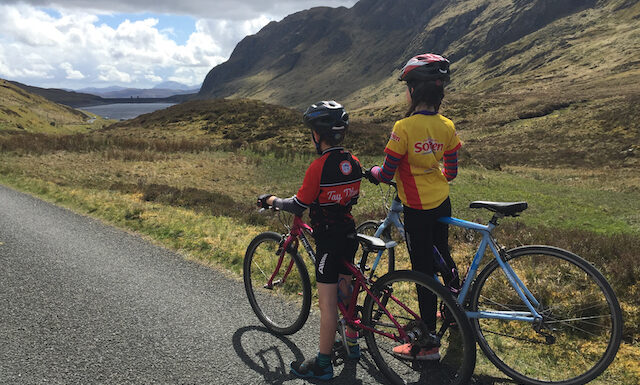 A young boy and girl stand astride their bikes looking south towards Lochan na Lairige, just below the summit of Ben Lawers. The girl is wearing a Soreen Malt Loaf cycling jersey and the boy is wearing a Tay Titans Junior Cycling Club jersey.