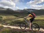 A young girl cycles on a mountain trail through the Cairngorms with the northern corries of Cairngorm behind her