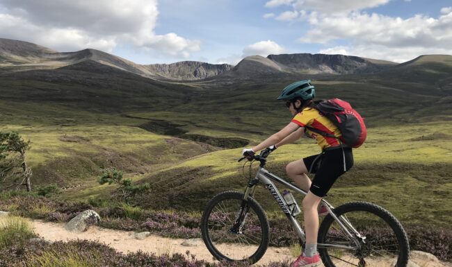 A young girl cycles on a mountain trail through the Cairngorms with the northern corries of Cairngorm behind her