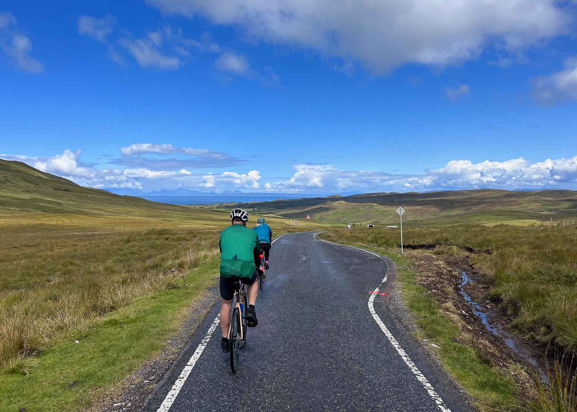 Two riders on a single track road below blue skies