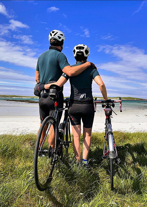 Cycling holidays in Scotland. A male and female cyclist stand with their bikes and an arm around each other on a grassy patch overlooking a white-sand beach on North Uist